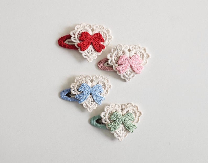 Pu&ograve; includere: Quattro fermagli per capelli a forma di cuore con dettagli in pizzo all'uncinetto e fiocchi. I fermagli presentano fiocchi rossi, rosa, blu e verdi, ciascuno su una forcina abbinata. Il pizzo &egrave; bianco, su sfondo bianco.