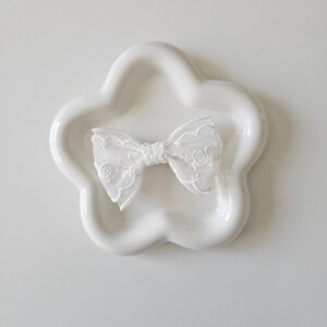 May include: A white lace bow on a white ceramic flower-shaped dish.