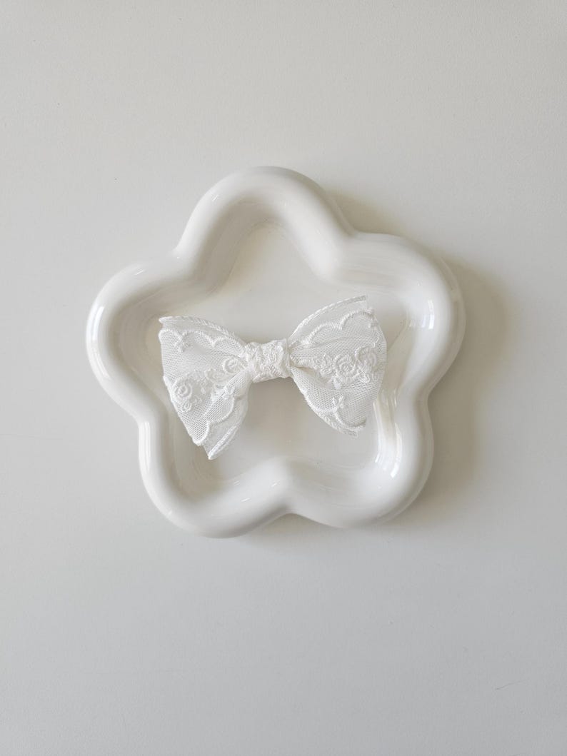 May include: A white lace bow on a white ceramic flower-shaped dish.