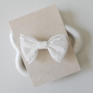 May include: A white lace bow with a delicate floral pattern. The bow is on a beige card with a repeating pattern.