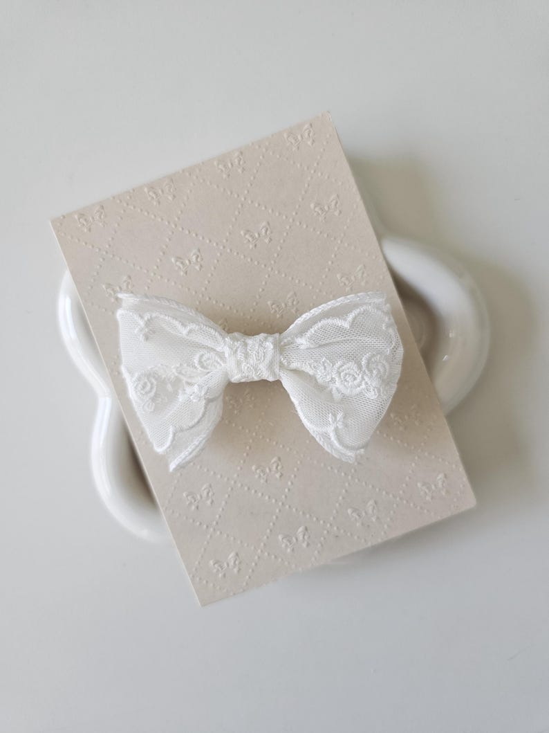 May include: A white lace bow with a delicate floral pattern. The bow is on a beige card with a repeating pattern.