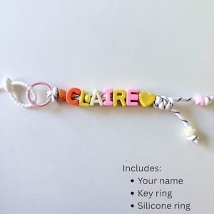 Op de afbeelding: Een gepersonaliseerde sleutelhanger met de naam CLAIRE gespeld in kleurrijke letterkralen, inclusief oranje, gele en roze tinten. Het ontwerp bevat een gele hartvormige kraal, een roze siliconen ring en een wit koord met zwarte spikkels, eindigend in stervormige kralen.