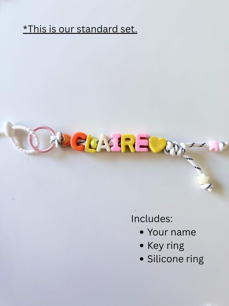 Op de afbeelding: Een gepersonaliseerde sleutelhanger met de naam CLAIRE gespeld in kleurrijke letterkralen, inclusief oranje, gele en roze tinten. Het ontwerp bevat een gele hartvormige kraal, een roze siliconen ring en een wit koord met zwarte spikkels, eindigend in stervormige kralen.