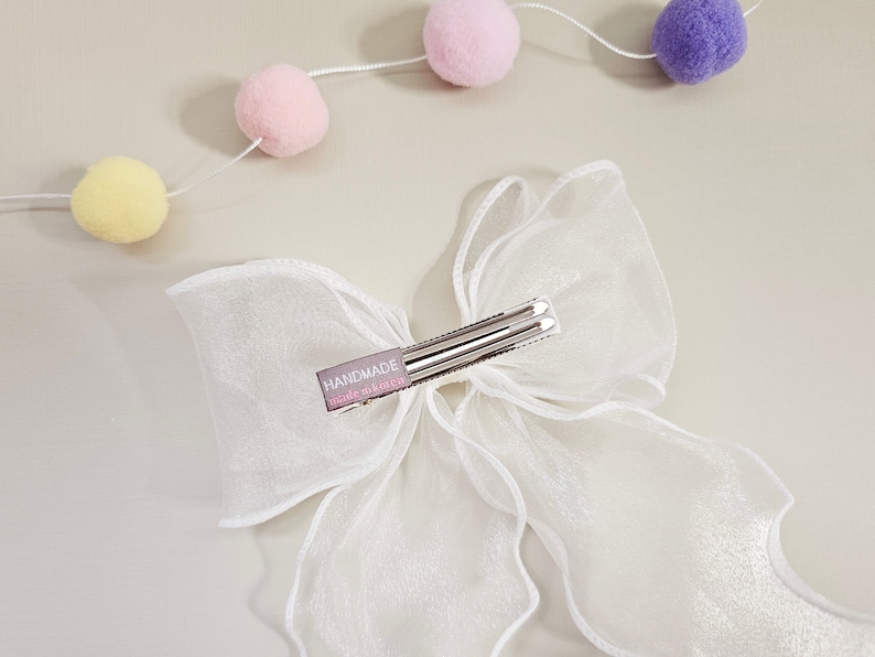 May include: A white sheer ribbon bow hair clip with a silver clip and a small tag that says "HANDMADE" with the words "made with love" underneath.