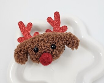 Puede incluir: Un reno de peluche marrón con cuernos de purpurina roja y una nariz de fieltro roja.