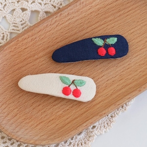 May include: Two embroidered hair clips with a cherry design. One clip is navy blue, the other is off-white. Each clip features a pair of red cherries with green leaves. The clips are displayed on a wooden tray.