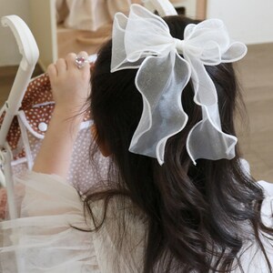 May include: A young girl with long dark hair wearing a white dress with a large white bow in her hair. She is holding a brown and white polka dot stroller.