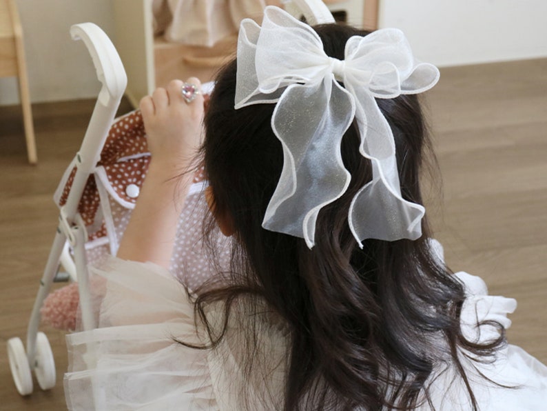 May include: A young girl with long dark hair wearing a white dress with a large white bow in her hair. She is holding a brown and white polka dot stroller.