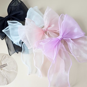 May include: Four sheer ribbon bows in black, white, pink, and purple. The bows are tied with a simple knot and have a ruffled edge. The bows are arranged in a semi-circle on a light beige background.