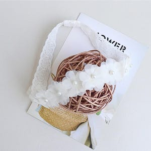 May include: White headband with a row of white fabric flowers and pearl accents. The headband is made of lace and is tied at the back.