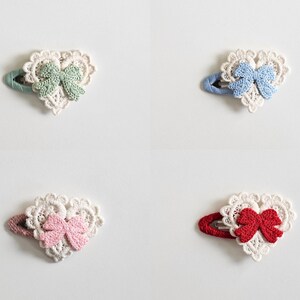 Pu&ograve; includere: Quattro fermagli per capelli a forma di cuore con dettagli in pizzo e un fiocco. Ogni fermaglio presenta un fiocco di colore diverso: verde, blu, rosa e rosso. I fermagli sono su sfondo bianco.