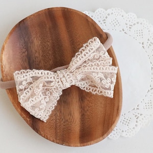 Puede incluir: Un lazo de encaje beige sujeto a una diadema elástica fina a juego. El lazo presenta un intrincado patrón floral con bordes festoneados, confeccionado con un tejido delicado y semitransparente que crea una apariencia texturizada y en capas.