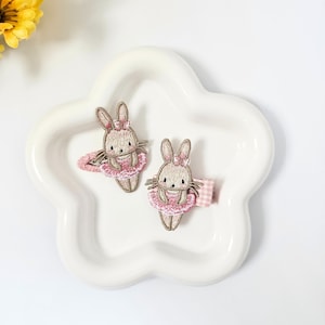 Op de afbeelding: Twee bruine geborduurde konijnenhaarclips met roze tutu's en witte geruite lintaccenten. De clips liggen op een wit stervormig schaaltje.