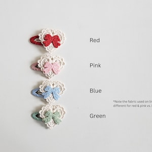 Pu&ograve; includere: Quattro fermagli per capelli a forma di cuore rossi, rosa, blu e verdi. Ogni fermaglio presenta un cuore di pizzo bianco con un fiocco abbinato. Il testo indica i colori.