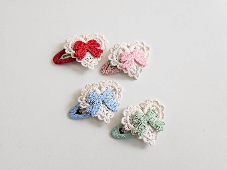 Pu&ograve; includere: Quattro fermagli per capelli a forma di cuore con pizzo all'uncinetto e fiocchi. I fiocchi sono rossi, rosa, blu e verdi. Le clip sono in vari colori, tra cui rosso, rosa, blu e verde. Gli accessori per capelli sono su sfondo bianco.