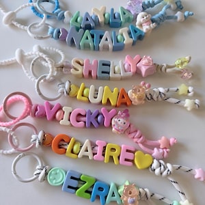 Op de afbeelding: Zes kleurrijke kralen-sleutelhangers met namen in plastic letterblokjes, waaronder Layla, Natalia, Shelly, Luna, Vicky, Claire en Ezra. Elke sleutelhanger bevat een witte of gekleurde ring, een nylon koord en diverse bedels zoals sterren, hartjes en dieren.