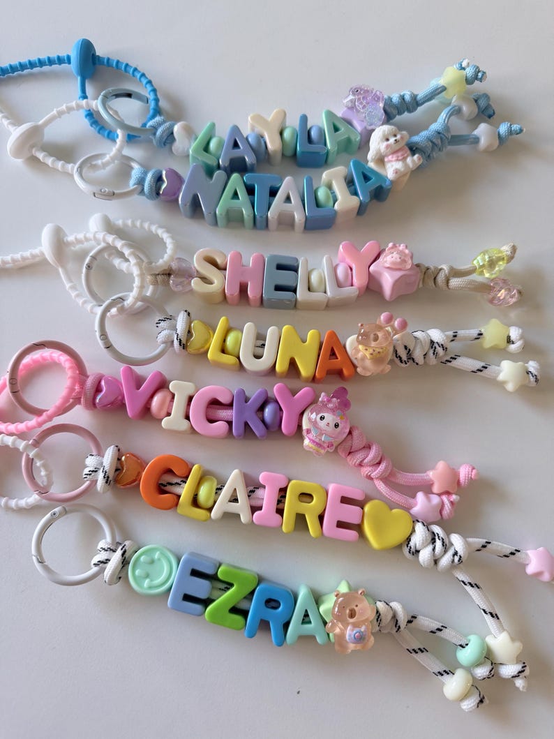 Op de afbeelding: Zes kleurrijke kralen-sleutelhangers met namen in plastic letterblokjes, waaronder Layla, Natalia, Shelly, Luna, Vicky, Claire en Ezra. Elke sleutelhanger bevat een witte of gekleurde ring, een nylon koord en diverse bedels zoals sterren, hartjes en dieren.