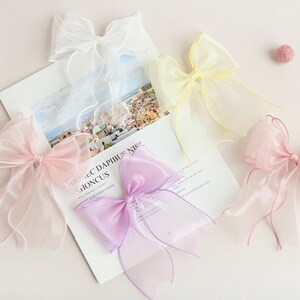 May include: Four sheer ribbon bows in different colours: white, yellow, pink, and purple. The bows are tied with long tails and are arranged on a white surface.