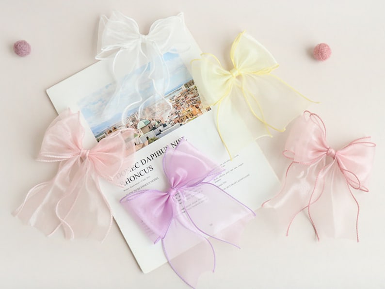 May include: Four sheer ribbon bows in different colours: white, yellow, pink, and purple. The bows are tied with long tails and are arranged on a white surface.