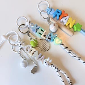 Op de afbeelding: Drie gepersonaliseerde sleutelhangers met witte koorden en metalen uiteinden. E&eacute;n toont de naam GINA met een golfclub en balbedel. Een andere toont KELLY met een tennisracket en balbedel. De derde toont RYAN met een honkbalknuppel en balbedel.