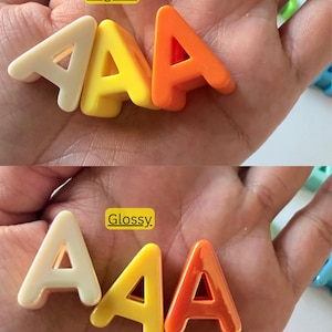 Op de afbeelding: Drie letter A-vormen in cr&egrave;me, geel en oranje. Het bovenste gedeelte toont de letters met een normale afwerking, terwijl het onderste gedeelte dezelfde letters met een glanzende afwerking laat zien. Elke letter is een solide, driedimensionaal object.