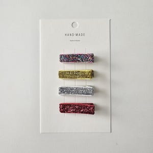 Könnte beinhalten: Satz mit vier Glitzer-Haarklammern in Rot, Gold, Silber und mehrfarbigem Glitzer. Die Klammern werden auf einer weißen Karte mit dem Text "HAND-MADE Made in Korea" präsentiert.