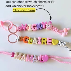 Op de afbeelding: Drie gepersonaliseerde kralenkoorden met kleurrijke letterkralen die VICKY, CLAIRE en MAMA spellen. De koorden bevatten roze en witte touwtjes, hartvormige kralen en een kleine figuurbedel. Tekst: You can choose which charm or I&rsquo;ll add whichever looks best :) *Add-on charm.
