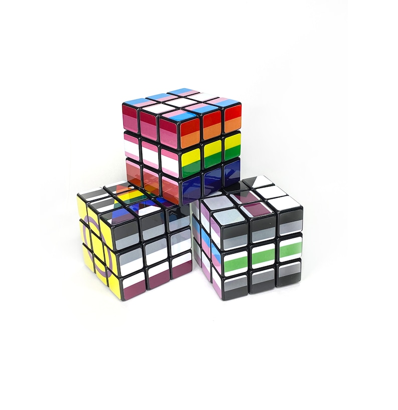 May include: Three 3x3 Rubik's Cubes with different pride flag designs. The top cube is a rainbow flag, the left cube is a bisexual flag, and the right cube is a transgender flag.