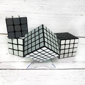 Peut inclure: Trois cubes de Rubik 3x3 noirs et blancs, dont un sur un support en plastique transparent. Le cube sur le support a un motif gris et blanc. Les deux autres cubes ont un motif noir et blanc.