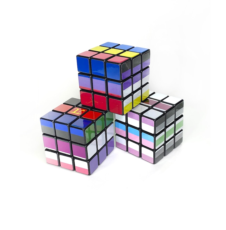 May include: Three 3x3 Rubik's Cubes with different flag designs. The top cube has a blue, yellow, and black design. The middle cube has a red, yellow, and blue design. The bottom cube has a purple, blue, white, and green design.