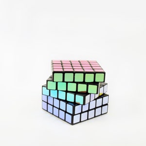 以下が含まれることがあります： 異なる色の面を持つ3つのルービックキューブの積み重ね。上のキューブはピンク、真ん中のキューブはグリーン、下のキューブはブルーです。