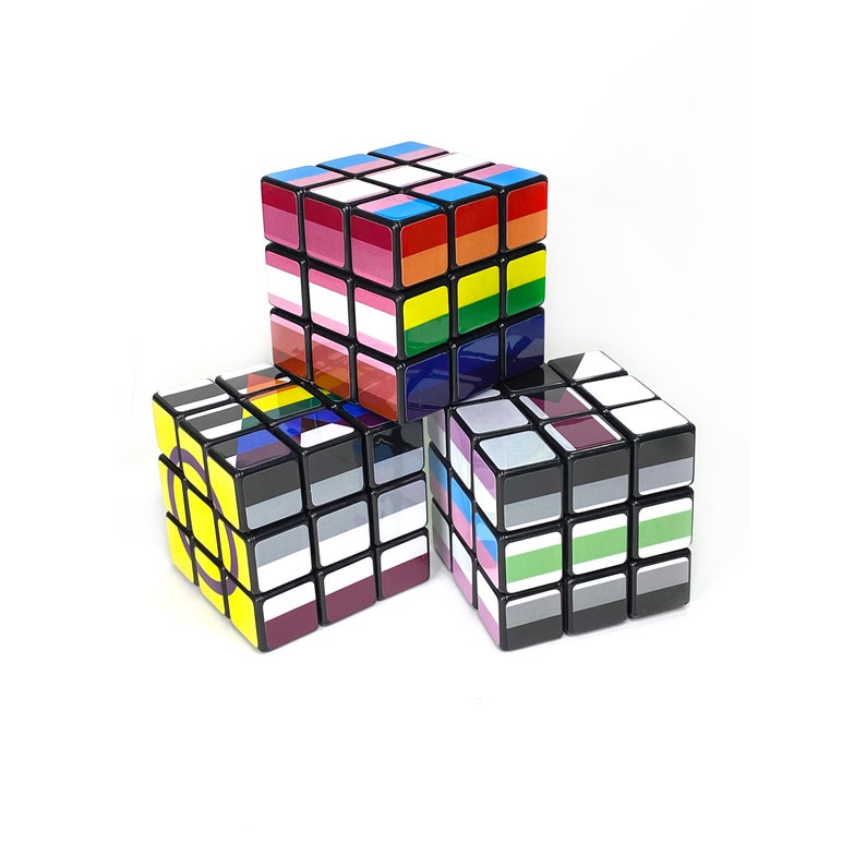 May include: Three 3x3 Rubik's Cubes with different pride flag designs. The top cube has a rainbow flag design, the left cube has a bisexual flag design, and the right cube has a transgender flag design.