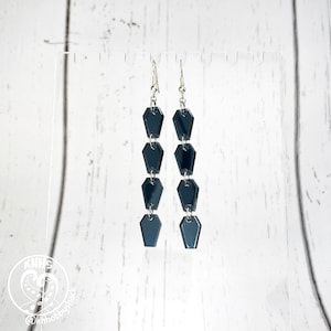 Peut inclure: Une paire de longues boucles d'oreilles pendantes en forme de cercueil noires avec des crochets argentés.