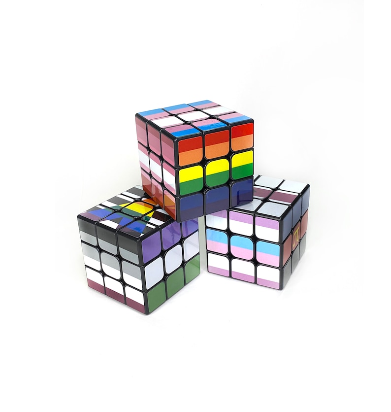 May include: Three 3x3 Rubik's Cubes with different pride flag designs. The top cube is a rainbow flag, the middle cube is a bisexual flag, and the bottom cube is a transgender flag.