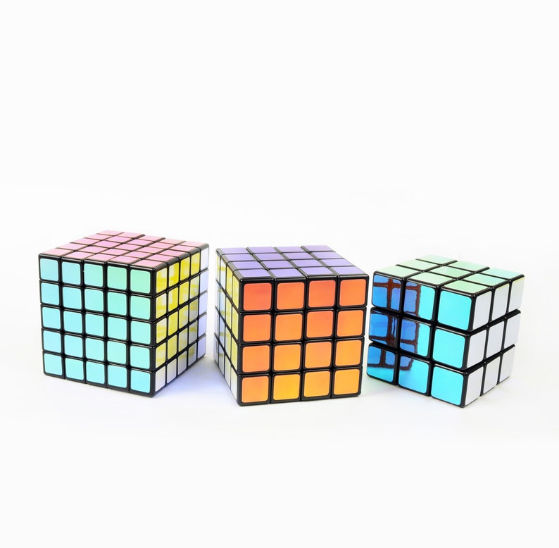 以下が含まれることがあります： 3x3のルービックキューブが3つ、それぞれ異なる色をしています。左側のキューブは青、緑、黄色の組み合わせです。中央のキューブはオレンジ、紫、白の組み合わせです。右側のキューブは青、緑、白の組み合わせです。
