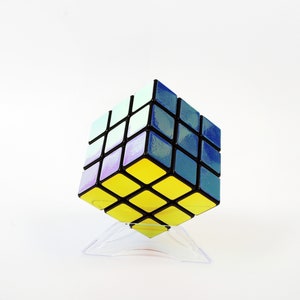 以下が含まれることがあります： 青、紫、白の虹色の仕上げが施された黒と黄色の3x3ルービックキューブパズル。キューブは透明なプラスチックスタンドの上に置かれています。