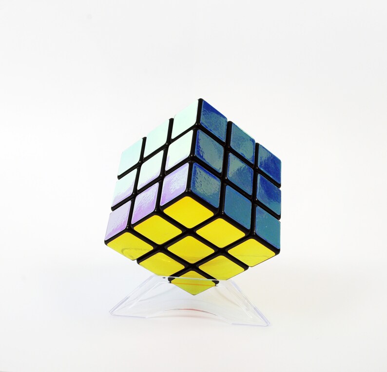 以下が含まれることがあります： 青、紫、白の虹色の仕上げが施された黒と黄色の3x3ルービックキューブパズル。キューブは透明なプラスチックスタンドの上に置かれています。