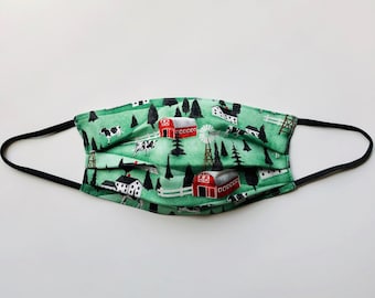 May include: A green fabric face mask with a black elastic band. The mask features a repeating pattern of red barns, white houses, black and white cows, and green trees on a green background.