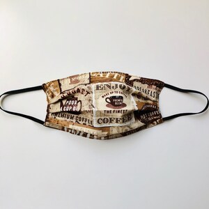 May include: A beige and brown fabric face mask with a coffee theme. The mask features text that says "Enjoy a Spiced Coffee", "Pour a Cup of", "Premium Coffee", "Wake Up to Coffee", "The Finest Coffee", "Hot Coffee", and "Breakfast".