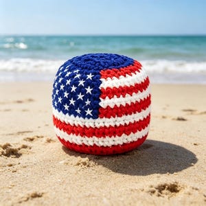 May include: A crocheted ball featuring the design of the American flag, with blue, white, and red stripes and stars. The ball is resting on a sandy beach with the ocean in the background.