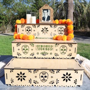 May include: A three-tiered wooden altar for Día de los Muertos, featuring laser-cut skull and floral designs. The altar is decorated with orange and yellow marigolds, candles, and a framed portrait. The text "Día de los Muertos" is visible.