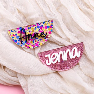 May include: Two half-circle acrylic name tags with glitter and confetti designs. The top tag is multi-colored with the name "Anne" in black lettering. The bottom tag is pink with the name "Jenna" in white lettering.