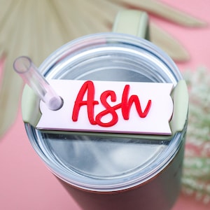 Puede incluir: Un vaso de acero inoxidable con tapa transparente y una pajita. Una etiqueta blanca con el nombre "Ash" escrito en cursiva roja está unida a la tapa.