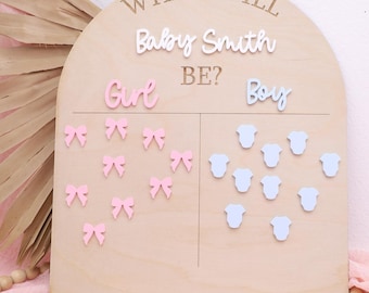 May include: Wooden gender reveal board with the text "WHAT WILL BABY SMITH BE?" in white. The board has sections for "Girl" and "Boy", with pink bows and blue onesies to indicate the baby's gender. A decorative item for a baby shower.