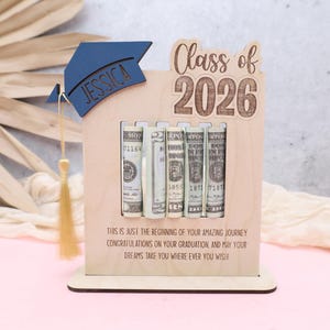 May include: Wooden graduation gift box with the text "Class of 2026". The box has a navy blue graduation cap with the name "Jessica" and holds rolled-up bills. The bottom of the box has the text "This is just the beginning of your amazing journey..."