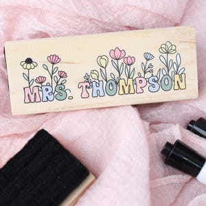 May include: A wooden dry erase board eraser with the name "MRS. THOMPSON" in pastel colors, decorated with floral illustrations. The eraser is on a pink fabric surface, with two black dry erase markers.