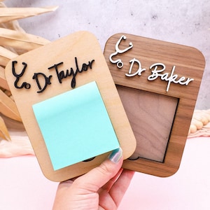 May include: Two wooden note holders with a stethoscope symbol and the text "& Dr. Taylor" and "& Dr. Baker". One holder has a light blue sticky note attached.