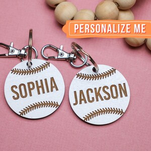 Puede incluir: Dos llaveros de madera blanca con forma de pelota de béisbol con anillas de metal plateadas. Los llaveros están personalizados con los nombres "Sophia" y "Jackson".