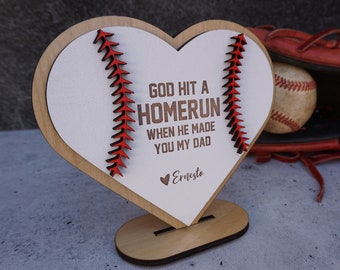 May include: A wooden heart-shaped sign with a baseball stitch design and the text "God hit a homerun when he made you my dad" with the name "Ernesto" at the bottom. The sign is on a small wooden stand.