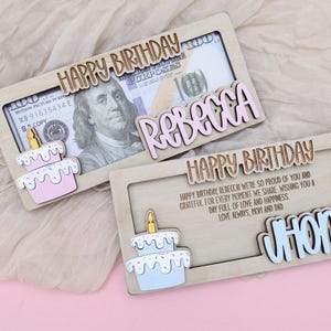 Puede incluir: Dos soportes de tarjetas de cumplea&ntilde;os de madera con una ranura para dinero en efectivo. La tarjeta superior tiene un pastel rosa y blanco con una vela amarilla y el texto "HAPPY BIRTHDAY REBECCA". La tarjeta inferior tiene un pastel azul y blanco con una vela amarilla y el texto "HAPPY BIRTHDAY CHONMY".